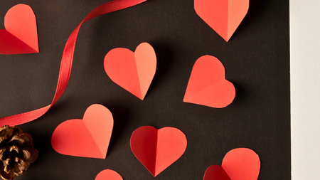 A collection of red paper hearts and a red ribbon are scattered across a dark, textured background,...