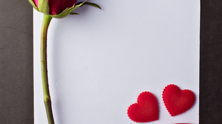 A single red rose lies beside two small red hearts on a white surface, evoking romance and affection.の写真素材