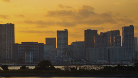 A city skyline is dramatically silhouetted against a vibrant, cloudy golden sunset, with a hint of water in the foreground.の写真素材