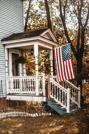 A cozy porch of a small house with an American flag hanging outside during autumn.