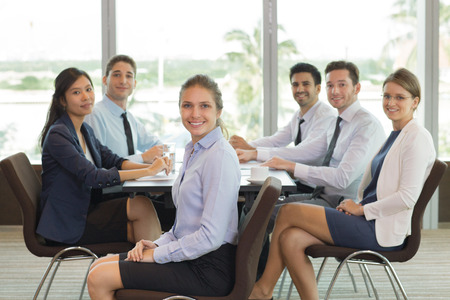 Young Caucasian business leader sitting at top of office desk with her business team on background. They are looking at camera and smilingの写真素材