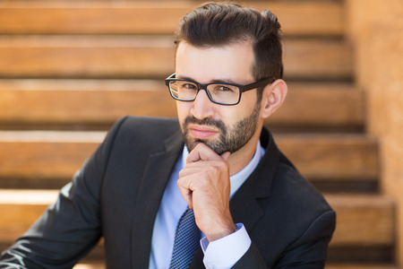 Portrait of young businessman sitting outside office, holding his chin with hand and having confident look at cameraの写真素材