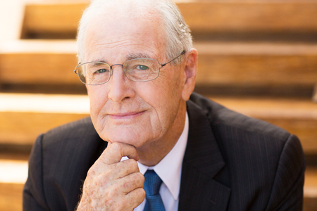 Portrait of mature grey-haired businessman sitting outside office, holding his chin with hand and smiling at cameraの写真素材