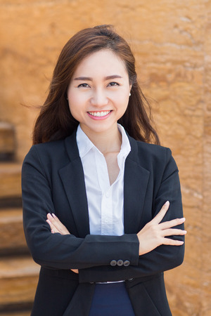 Portrait of young Asian businesswoman standing with crossed arms outside office and smiling at cameraの写真素材