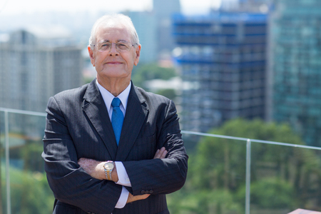 Portrait of senior businessman standing with crossed arms on rooftop of office building and looking at cameraの写真素材