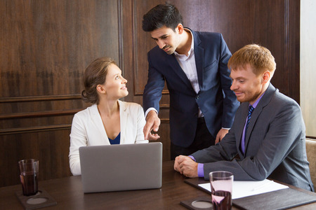 Young businessman explaining something and pointing to laptop screen while standing between male and female colleagues sitting at table and listening to himの写真素材