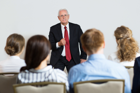 Senior businessman leaning on table, gesturing and telling something emotionally to sitting audience of four people. Rear view of audience.の写真素材