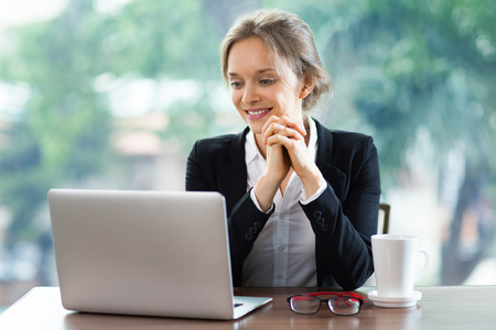 Smiling young attractive businesswoman sitting at table while looking at laptop screen with her hands clasped. Front view with big window and blurry green view outside in background.の写真素材