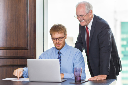 Young businessman sitting at table, pointing to paper with diagram and telling something to smiling senior boss standing behind. They are looking at laptop screen.の写真素材