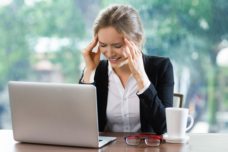 Young businesswoman sitting at table in front of laptop while closing eyes tight and touching temples. Front view with big window and blurry green view outside in background.の写真素材