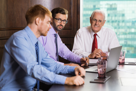 Senior and two young businessmen looking at laptop screen and discussing project while sitting at table in meeting room with window and blurry building view in backgroundの写真素材