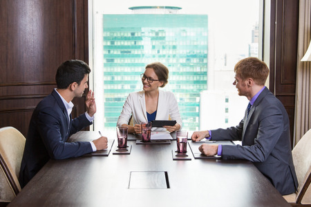 Smiling young businesswoman sitting at head of big table in meeting room with big window in background. Man sitting on her right is explaining something. Man sitting on her left is looking at them.の写真素材