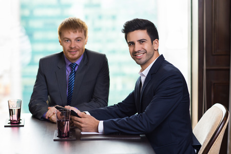 Two young handsome businessmen smiling and looking at camera while sitting at table in meeting room with big window and blurry city view in background. Farther man is slightly blurred.の写真素材