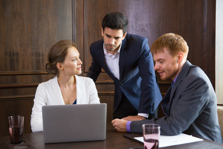 Young businessman thinking while looking at laptop screen and standing between male and female colleagues sitting at table. Woman is looking at sitting man who is telling something.の写真素材