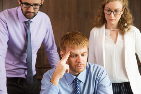 Close up portrait of three business people looking at something out of view and thinking. One businessman is sitting and leaning head on hand. Other male and female colleagues are standing behind.の写真素材