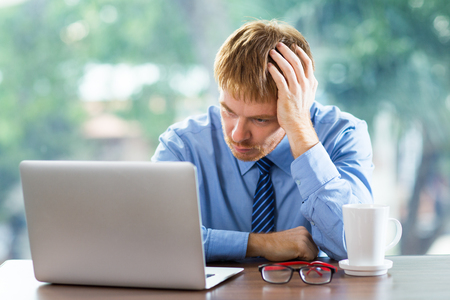 Thoughtful young businessman sitting at table and looking at laptop screen while leaning his head on hand. Front view with big window and blurry green view outside in background.の写真素材