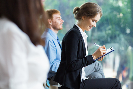 Smiling young businesswoman sitting and making notes with blurry businessman in background. Side view with big window and blurry green view outside in background.の写真素材