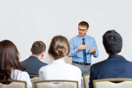 Standing young businessman gesturing and explaining something to sitting audience of four people. Rear view of audience.の写真素材