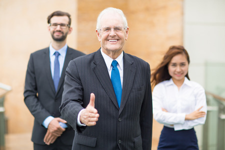 Smiling mature businessman standing on outside office and offering his hand to shake to business partner. His colleagues standing on backgroundの写真素材