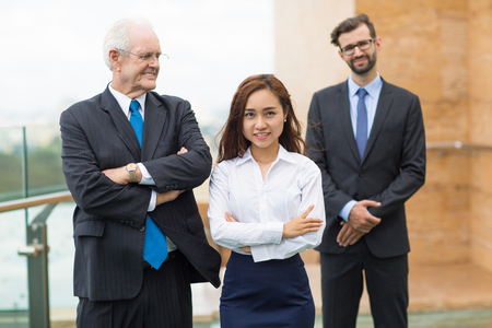 Successful mature businessman standing with crossed arms outside office and looking at his young assistant. Their male colleague standing on backgroundの写真素材