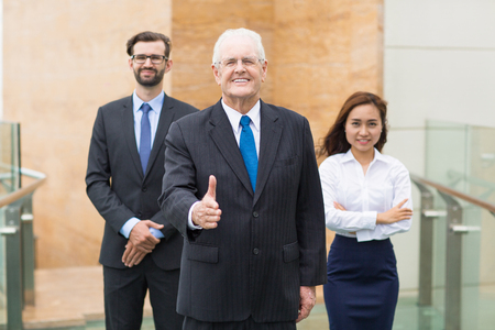 Successful mature businessman standing outside office and offering his hand to shake to partner. His colleagues standing on backgroundの写真素材