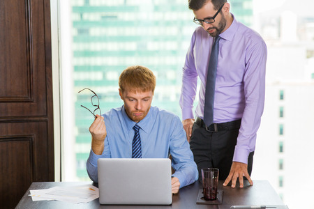 Young businessman standing behind colleague sitting at table with big window and blurry city view in background. They are looking at laptop screen and thinking.の写真素材