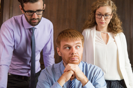 Close up portrait of three young business people thinking. One businessman is sitting, leaning his head on hands and looking aside. Other male and female colleagues are standing behind.の写真素材