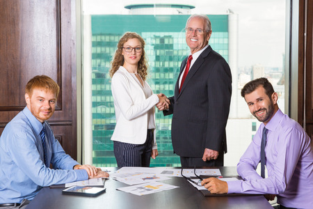 Senior business leader and young businesswoman standing at head of table and shaking hands. Another two businessmen are sitting at opposite sides of table. Everyone is smiling and looking at camera.の写真素材