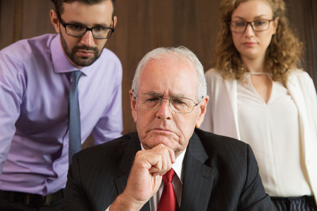 Close up portrait of pensive senior businessman leaning his chin on hand with young male and female colleagues in backgroundの写真素材