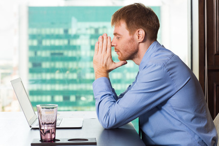 Young businessman sitting at table and looking at laptop screen while leaning his head on hands pressed together. Side view in front of big window with blurry city view outside.の写真素材
