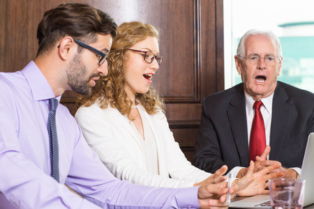 Senior businessman and young male and female colleagues looking at laptop screen with open mouths in astonishment while sitting at table in meeting roomの写真素材