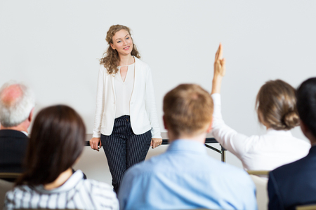 Smiling young businesswoman leaning on table and looking at woman from audience raising hand. Rear view of audience of five people.の写真素材