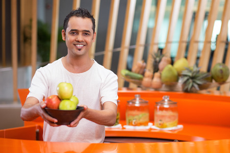 Portrait of smiling young man offering apples and standing behind bright fitness bar counter in gymの写真素材