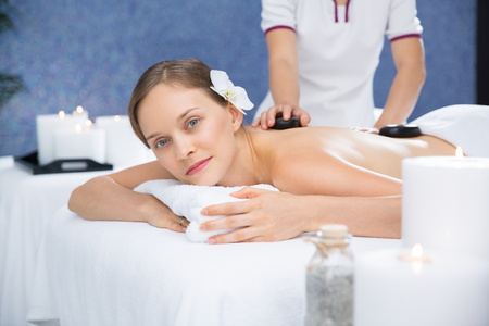 Content young woman lying on table, looking at camera and having stone massage therapy in spa salon room with lit candlesの写真素材