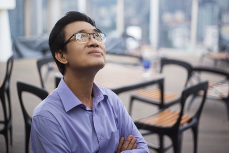Closeup portrait of thoughtful Asian senior man looking up with arms crossed in cafe with chairs and...