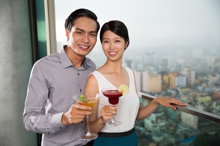 Smiling at camera young Asian woman and man holding glasses of cocktails and standing on balcony with blurred city view outsideの写真素材