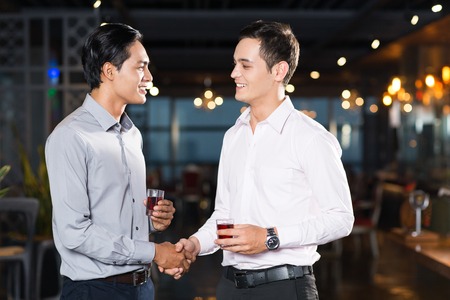 Two smiling young men shaking hands and holding glasses of wine at party in barの写真素材