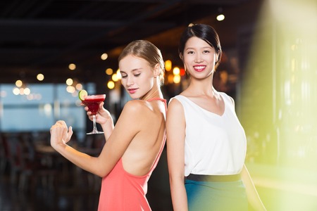 Two smiling young women standing back to back in nightclub with lights in background and ray of light in foreground. One woman is holding glass with cocktail and dancing.の写真素材
