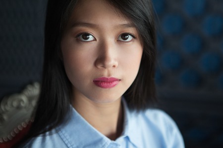 Closeup portrait of attractive young Asian woman with long black hair, wearing blue shirt, sitting in restaurant and looking at cameraの写真素材