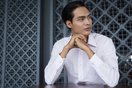 Portrait of serious young Asian businessman with his hands on chin sitting in restaurant, looking awayの写真素材