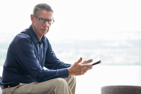 Portrait of frowning senior man sitting on chair, holding digital tablet, looking at camera seriouslyの写真素材