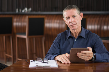 Portrait of thoughtful serious senior man using digital tablet, sitting at table in cafe, looking at cameraの写真素材