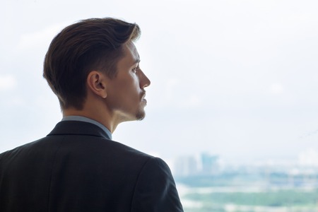 Profile portrait of serious young Caucasian businessman looking through window, thinking. Back view