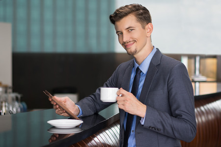 Closeup portrait of content middle-aged businessman looking at camera, holding tablet and cup, standing at cafe counterの写真素材