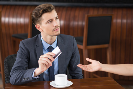 Portrait of serious middle-aged businessman sitting at cafe table and giving credit card to waiterの写真素材