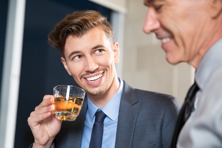 Close up of smiling young businessman holding cup of tea and looking at his senior colleague seen partly in foregroundの写真素材