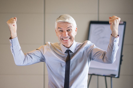 Closeup portrait of cheerful senior businessman raising hands and pumping fists with blurry flipchart in backgroundの写真素材