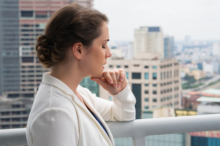Closeup of pensive adult businesswoman closing eyes and standing on roof top or balcony with blurry city view in background. Side view.の写真素材