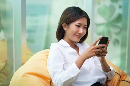 Smiling young Asian woman sitting in beanbag chair and using smartphone