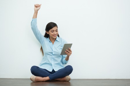Portrait of smiling young beautiful woman sitting on floor with crossed legs, using tablet and raising one hand with white wall in background. Front view.の写真素材